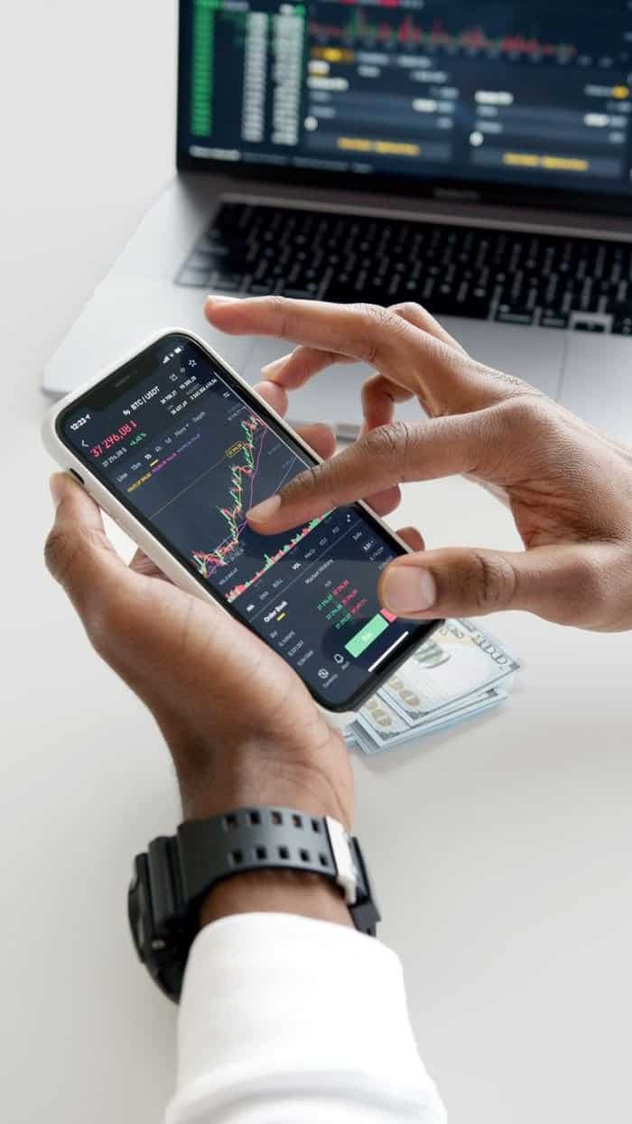 Home Hands using smartphone trading app with charts, dollar bills in background.